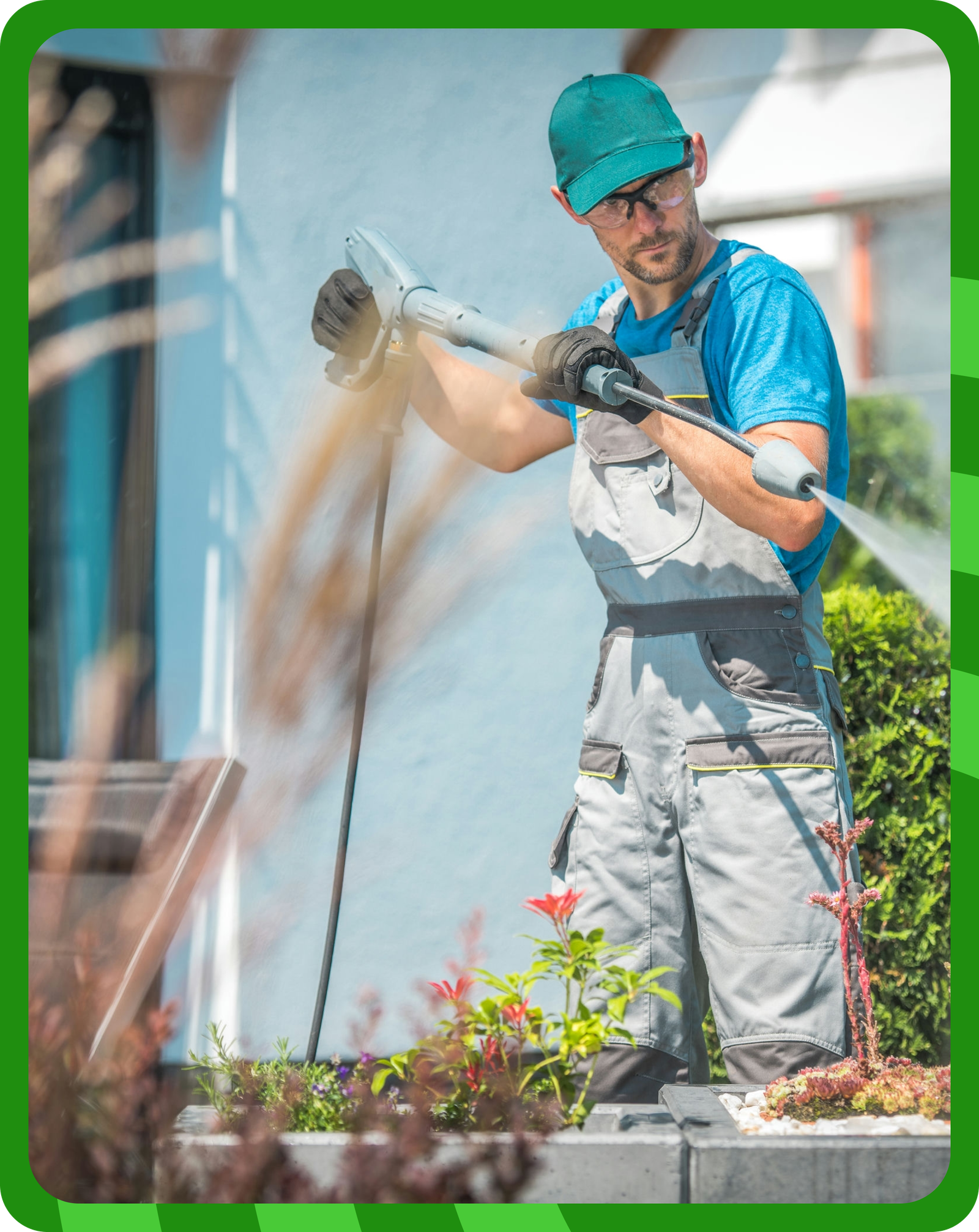 A professional worker wearing a blue cap, safety glasses, gloves, and gray overalls power washing an outdoor surface near a house. The worker is holding a high-pressure water hose, cleaning a paved area surrounded by plants and a garden.
